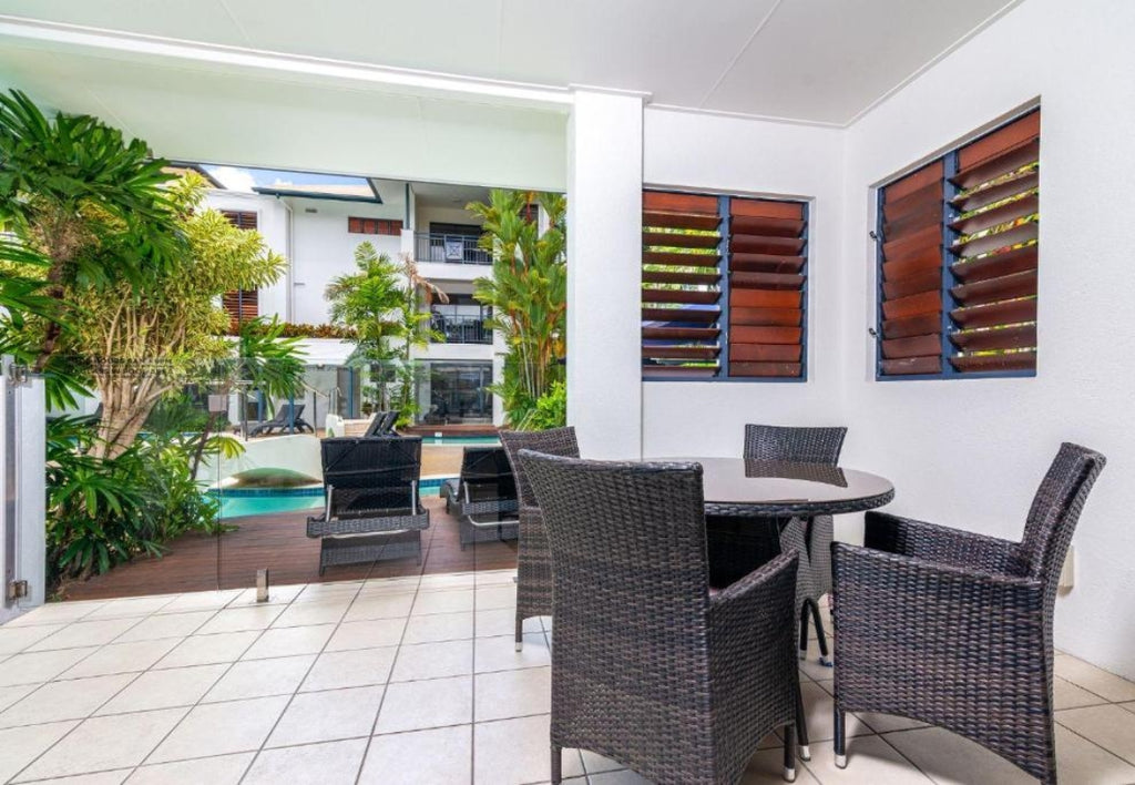 Outdoor patio area with wicker chairs and a dining table, pool visible through glass doors. Meridian Port Douglas Resort