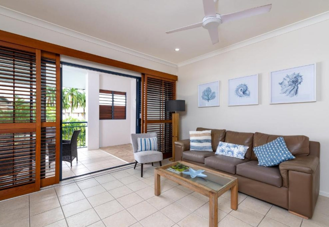 Living room with brown sofa, coffee table, and sliding glass doors leading to a balcony. Meridian Port Douglas Resort